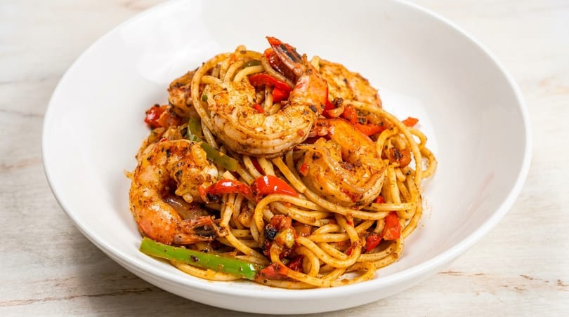 Spicy shrimp pasta with red and green bell peppers in a white bowl on a light wooden surface.
