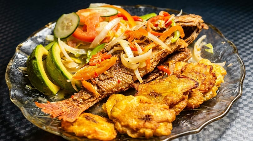Fried fish topped with onions and peppers, served with avocado slices, salad, and fried plantains.